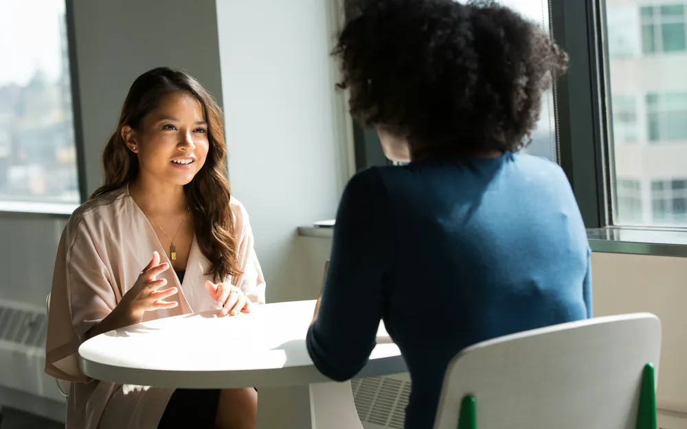 Two people talking across a small table by a window.
